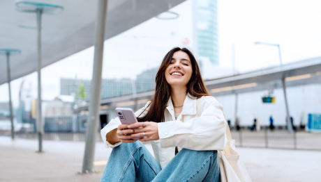 Frau mit braunen Haaren, weißer Bluse und hellblauen Jeans sitzt vor einem urbanen Hintergrund, lächelt in die Kamera und hält ein Handy mit beiden Händen.