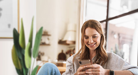Blonde Frau in Jeans sitzt zuhause und schaut in ihr Smartphone