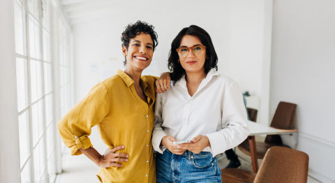 Zwei Frauen in Hemden stehen aneinander gelehnt im Büro und schauen in die Kamera