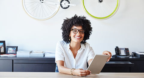 Frau mit schwarzen Locken und Brille in weißem Hemd hält Tablet in der Hand und schaut in die Kamera