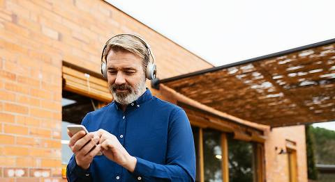 Mann in blauem Hemd steht vor einem Haus mit Kopfhörern auf und Smartphone in der Hand