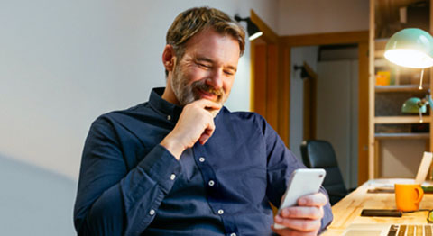 Mann in dunkelblauem Hemd sitzt am Schreibtisch und schaut in sein Smartphone