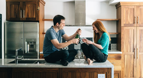 Frau und Mann sitzt in der Küche auf einem Tresen und öffnen eine Sektflasche zum Anstoßen