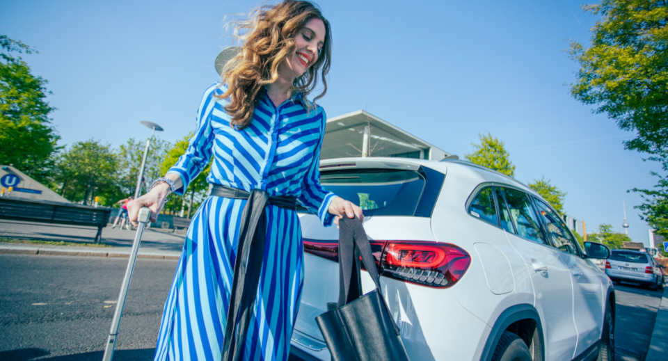 Lockige Frau in blau weiß gestreiftem Kleid läuft hinter einem weißen E-Auto