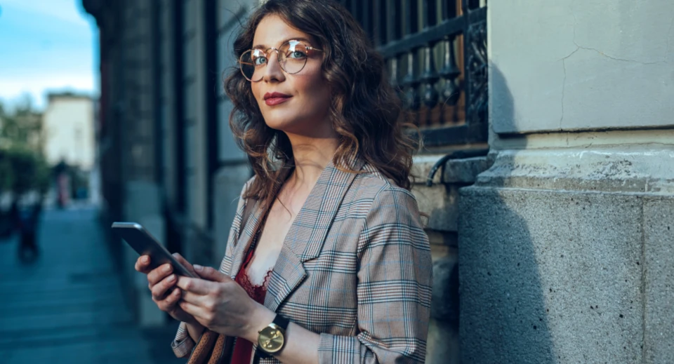 Lockige Frau mit Brille und Blazer hat ein Smartphone in der Hand und schaut in die Ferne