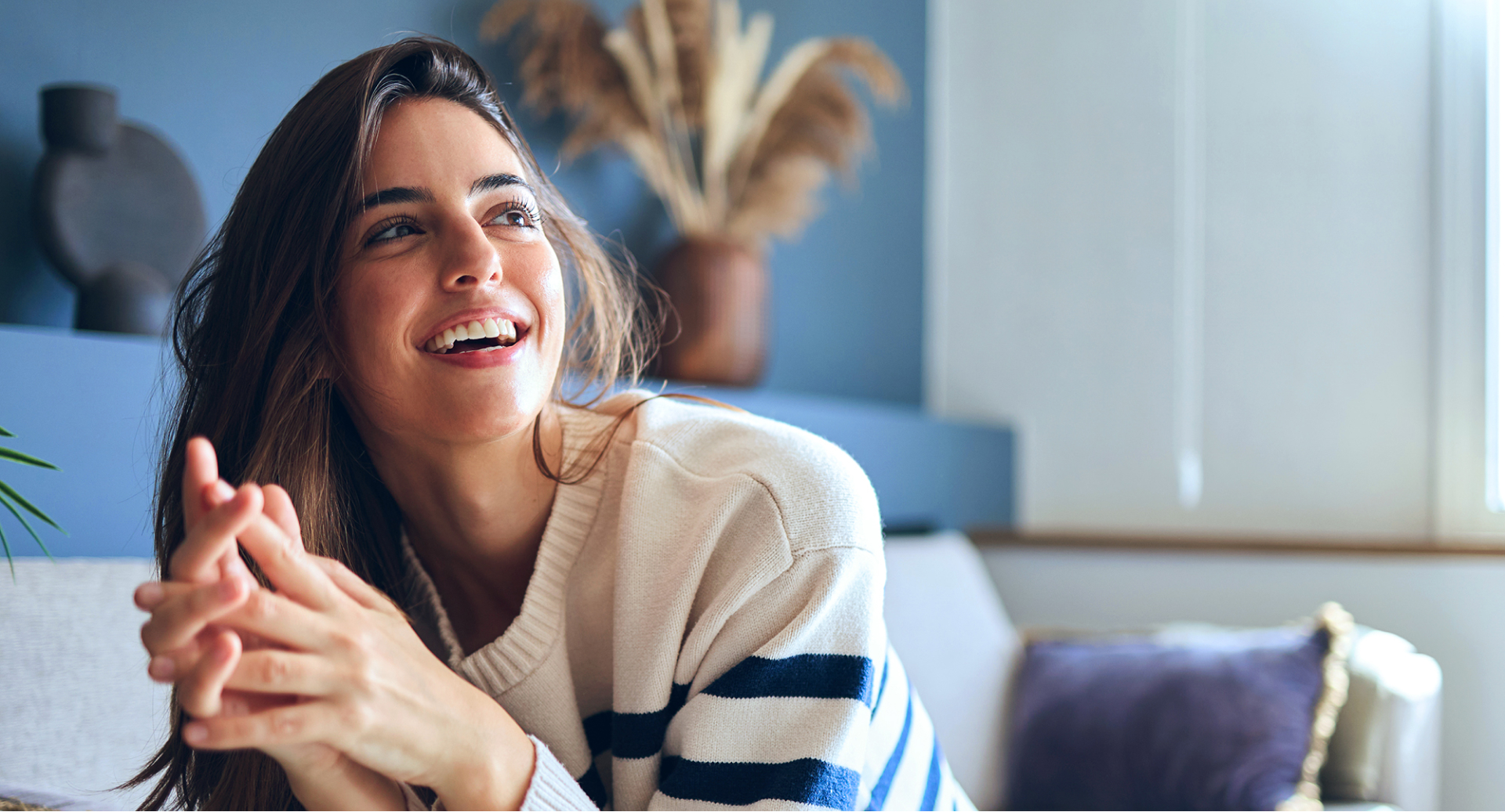 Dunkelhaarige Frau liegt lachend auf dem Bauch auf dem Sofa und schaut in die Ferne