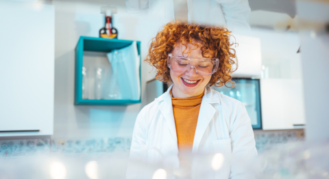 Frau mit leuchtend roten Locken und transparenter Brille steht mit weißem Kittel in einem Labor und blickt lächelnd nach untend 