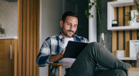 Mann im karierten Hemd sitzt mit überschlagenen Beinen in einem Stuhl, hält ein Tablet 