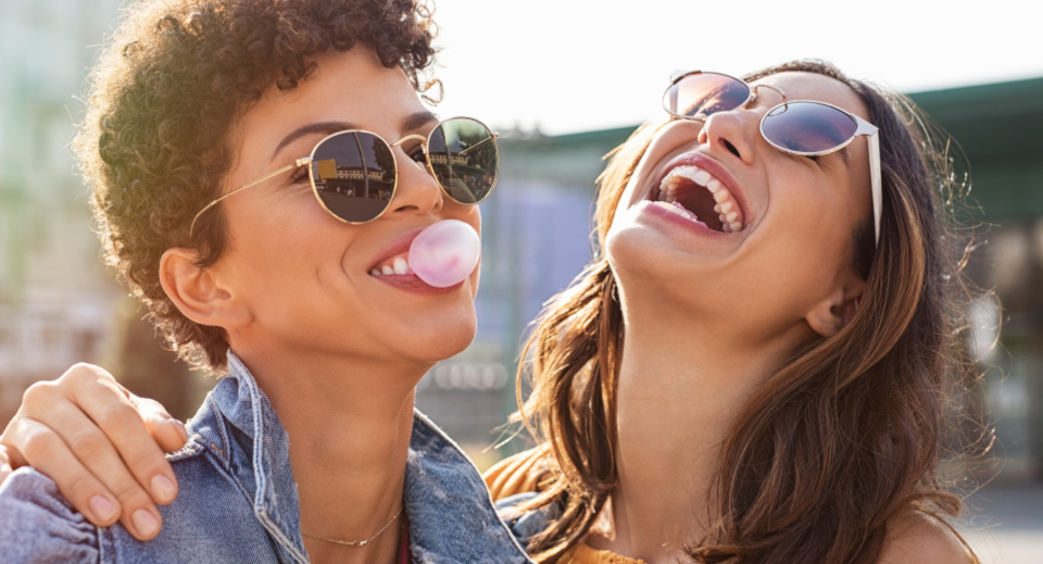 Zwei Frauen mit Sonnenbrille stehen draußen in der Abenddämmerung und lachen