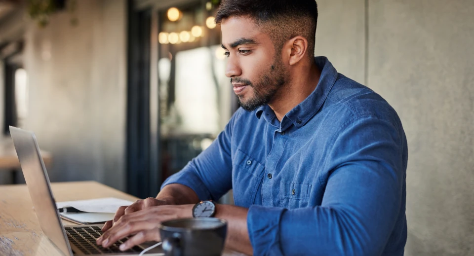 Dunkelhaariger Mann in Jeanshemd sitzt an einem Tisch und arbeitet am Laptop