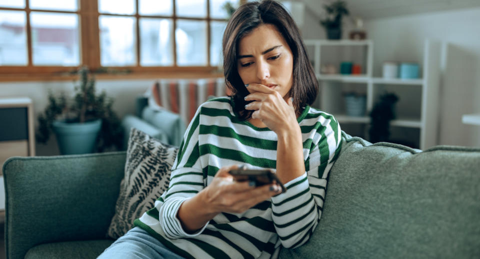 Dunkelhaarige Frau in grün weiß gestreiftem Pullover sitzt auf dem Sofa und schaut in ihr Smartphone