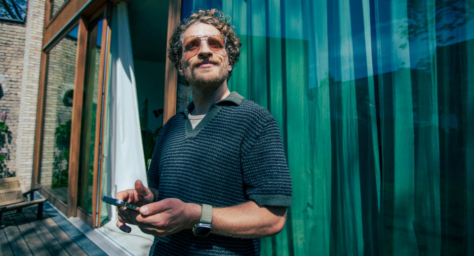 Lockiger Mann mit Sonnenbrille steht vor einem Haus mit bodentiefen Fenster und schaut in die Ferne