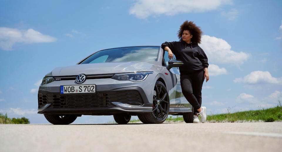 Frau mit braunen Locken steht angelehnt an einem grauen VW GTI
