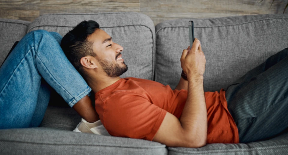 Dunkelhaariger Mann in orangenem T-Shirt liegt auf einem Sofa angelehnt an den Beinen einer anderen Person und schaut in sein Smartphone