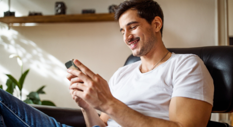 Mann mit kurzen braunen Haaren sitzt auf Sofa, lacht und schaut in sein Handy