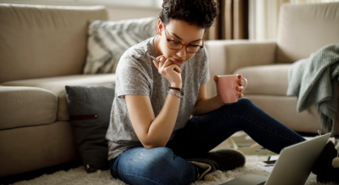 Frau mit kurzen braunen Haaaren und Brille sitzt auf dem Boden vorm Sofa und schaut auf Laptop