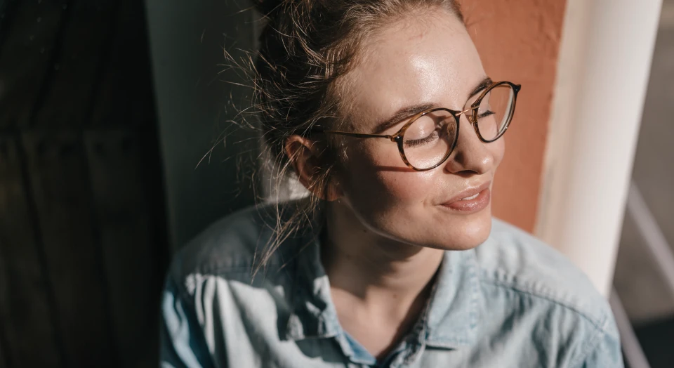 Frau mit Brille sitzt auf dem Boden vor dem Fenster mit geschlossenen Augen und genießt die Sonne