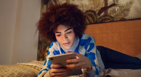 Frau mit braunem Afro liegt auf dem Bauch auf dem Bett und blickt auf ihr Tablet