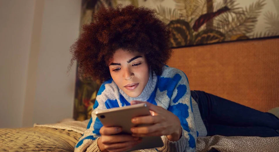 Frau mit Locken liegt auf dem Bauch auf ihrem Bett und schaut in ihr Tablet