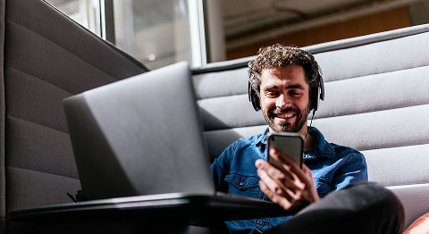 Lockiger Mann mit Kopfhörern sitzt in Arbeitsecke am Laptop und hält sein Smartphone in der Hand