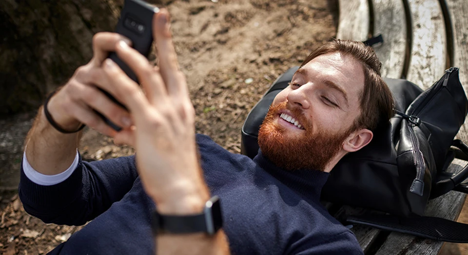 Mann mit Bart liegt im Freien an Baum angelehnt und grinst in sein Smartphone