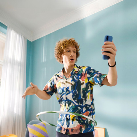 Eine Person befindet sich in einem hellen Raum mit türkisfarbener Wand und hält ein Smartphone in der rechten Hand. Die Person trägt ein bunt gemustertes Hemd und bewegt einen Hula-Hoop-Reifen um die Taille. Im Hintergrund stehen ein Sessel mit buntem Kissen und weitere Möbel.