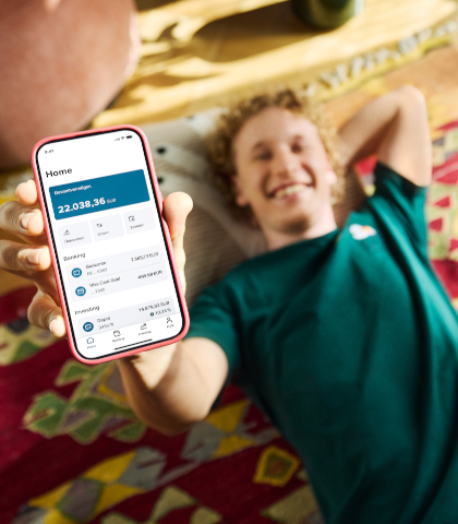 Eine Person liegt entspannt auf einem bunten Teppich und hält ein Smartphone in der Hand. Auf dem Bildschirm der Banking-App ist das Hauptmenü zu sehen. Der Gesamtvermögensstand beträgt 22.038,36 Euro. Unterteilt ist das Vermögen in zwei Bereiche: Banking mit einem Girokonto (7.385,73 Euro) und einer Visa Card Gold (499,98 Euro) sowie Investing mit einem Depot (14.879,32 Euro). Am unteren Bildschirmrand sind verschiedene Navigationssymbole sichtbar.