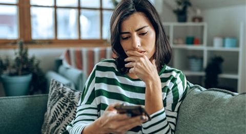 Frau mit langen braunen Haaren sitzt auf Sofa schaut skeptisch ins Handy