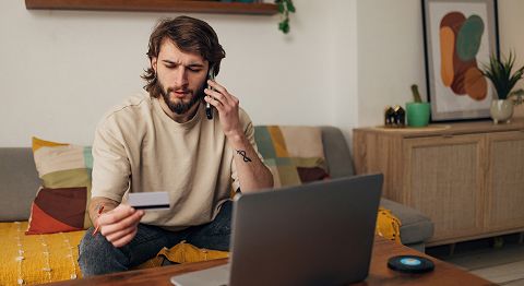 Mann mit langen braunen Haaren sitzt auf Sofa, schaut auf seine Kreditkarte und telefoniert