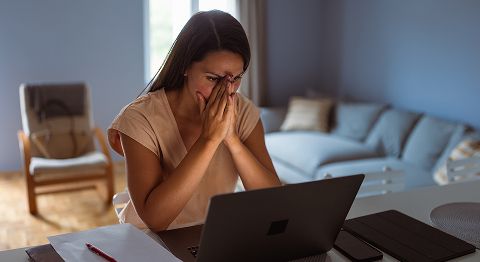 Frau mit langen braunen Haaren verschränkt Hände vorm Gesicht und sitzt am Schreibtisch vor Laptop
