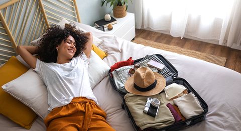 Frau mit langen gelockten braunen Haaren liegt auf Bett neben gepacktem Koffer