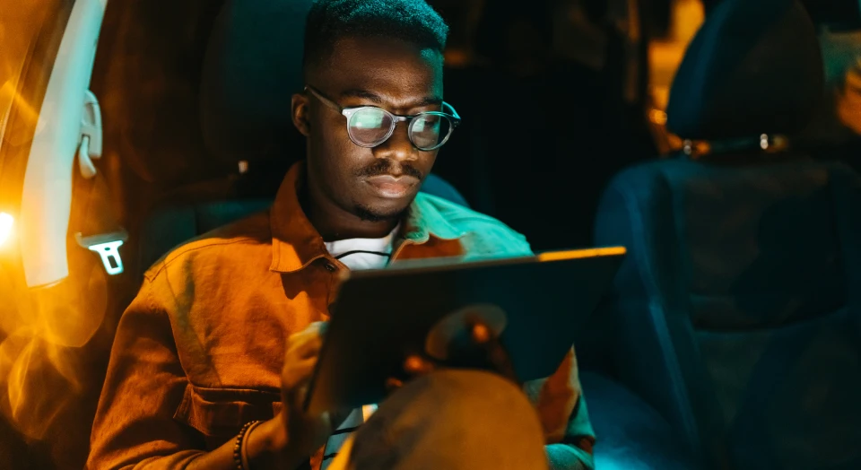 Mann mit Brille sitzt abends hinten in einem Auto und tippt etwas auf seinem Tablet