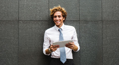 Lockiger Mann in weißem Hemd mit Krawatte hält Tablet in der Hand und lächelt in die Kamera