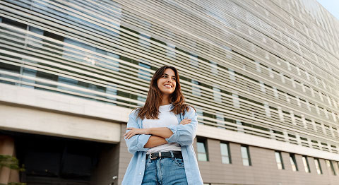 Braunhaarige Frau lächelt und steht mit verschränkten Armen vor einem Gebäude und schaut in die Ferne