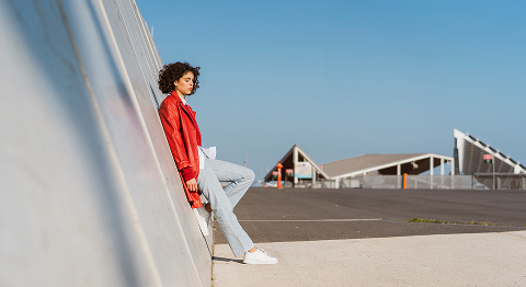 Dunkelhaarige Frau in roter Jacke lehnt an einer schrägen Gebäudewand