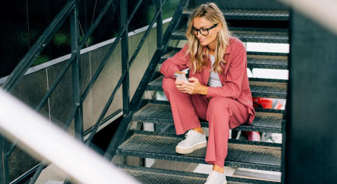 Frau in lässigem hellroten Anzug sitzt auf einer Treppe und schaut in ihr Smartphone