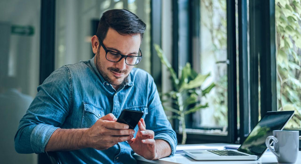 Mann mit schwarzer Brille und Jeanshemd sitzt am Schreibtisch und schaut in sein Smartphone