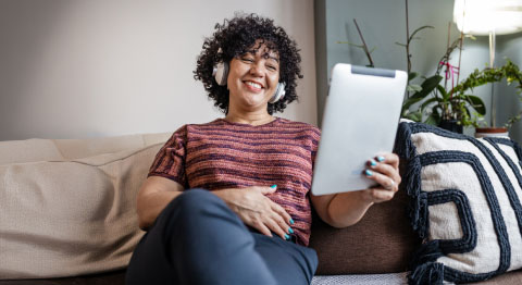 Lockige Frau mit Kopfhörern sitzt auf beigem Sofa und schaut in ihr Tablet