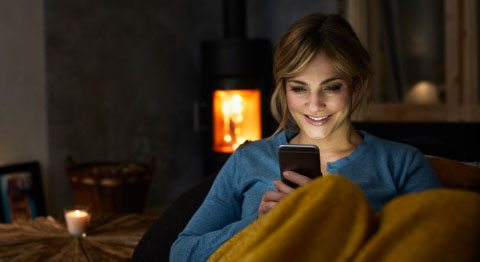 Blonde Frau sitzt abends zugedeckt auf ihrem Sofa und schaut in ihr Smartphone