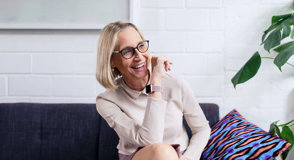 Blonde Frau mit schwarzer Brille sitzt auf dem Sofa und lacht zur Seite