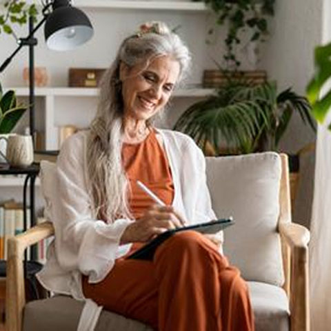 Frau mit grauen Haaren sitzt in ihrem Wohnzimmer auf einem Stuhl und macht sich Notizen am Tablet
