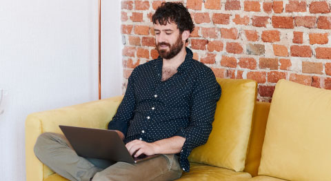Braunhaariger Mann in gepunktetem Hemd sitzt vor einer Backsteinwand auf dem Sofa und tippt auf seinem Laptop