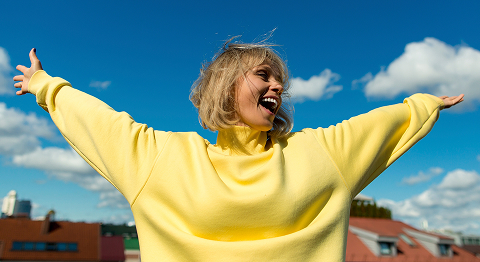 Frau mit gelben Pulli steht draußen, breitet  ihre Arme aus und lacht.