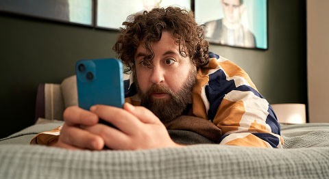 Lockiger Mann in buntem Hemd liegt auf dem Bauch in seinem Bett und schaut in sein blaues Smartphone