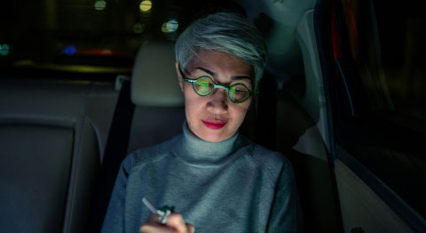 Frau mit grauen Haaren und Brille sitzt abends am Tablet in einem Auto und das Tablet spiegelt sich in ihren Brillengläsern