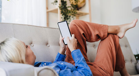 Blonde Frau in Jeansjacke liegt auf einem beigen Sofa und schaut in ihr Smartphone