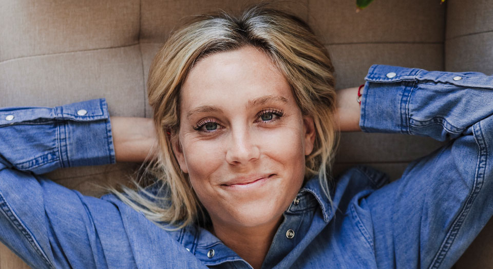 Blonde Frau in Jeanshemd liegt mit Armen hinter dem Kopf entspannt auf dem Sofa