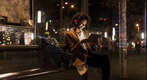 Lockige Frau mit Kopfhörern sitzt abends in einer Bushaltestelle und schaut auf ihr Tablet