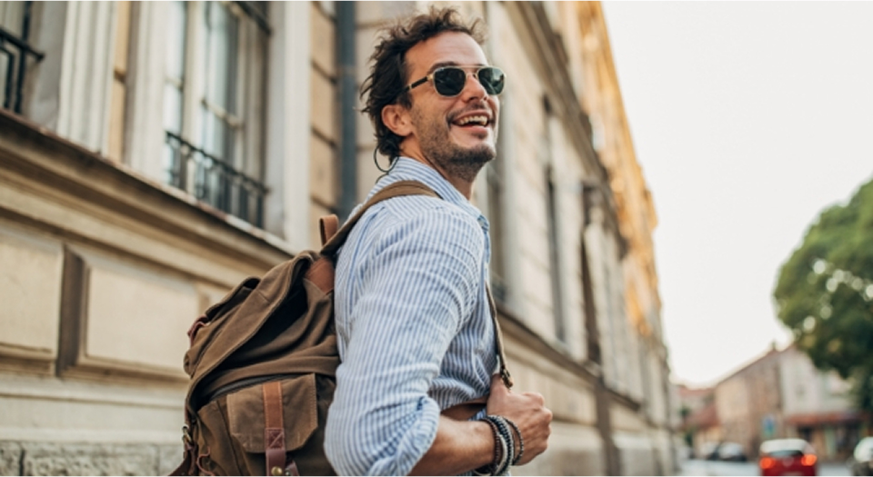 Mann mit Sonnenbrille in Jeanshemd mit Rucksack auf dem Rücken läuft draußen herum und schaut nach hinten