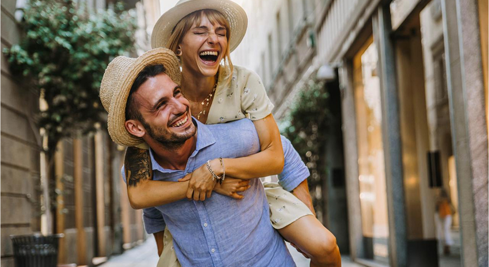 Mann mit Hut trägt Frau mit Hut auf den Schultern und beide lächeln